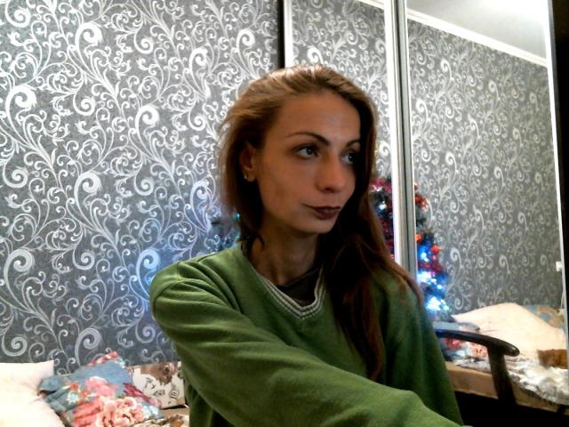 HotQueenAmelia with brown hair, green sweater, and dark lipstick, looking to the side with a slight smile, in a room with floral-patterned wallpaper and a small Christmas tree.