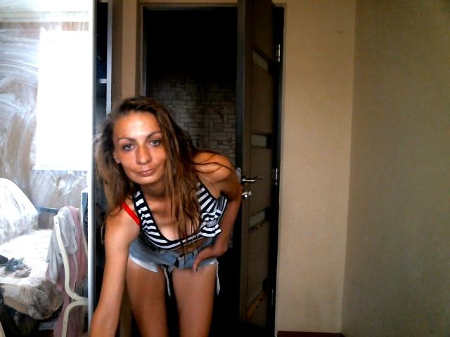 HotQueenAmelia with long brown hair, striped tank top, and denim shorts, bending forward, showing her small tits and red strap, in a dimly lit room.