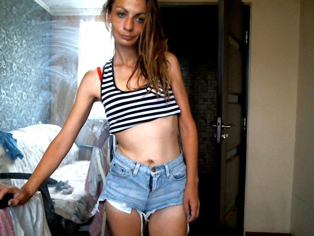 HotQueenAmelia stands in a room, wearing a black-and-white striped crop top and frayed denim shorts, showing a slim, toned midriff with small, perky tits and a subtle smile.
