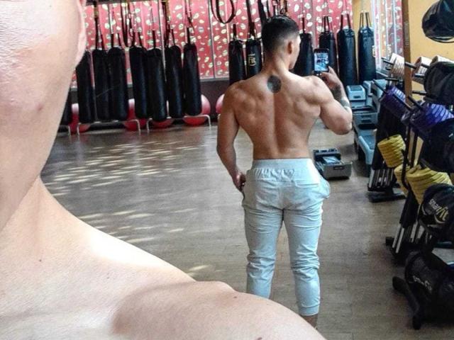 JuanJsmith is shirtless with a muscular back and a small tattoo, wearing gray pants, standing in a gym with a mirror showing his reflection.