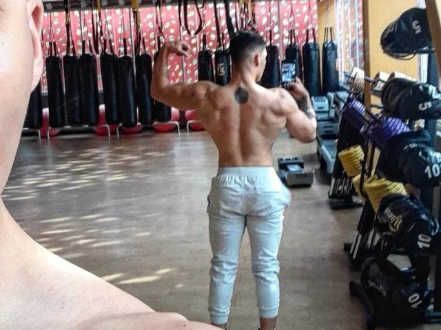 JuanJsmith, muscular and shirtless, flexes biceps in a gym, wearing gray pants, showing a black tattoo on his upper back, standing in front of weightlifting equipment.