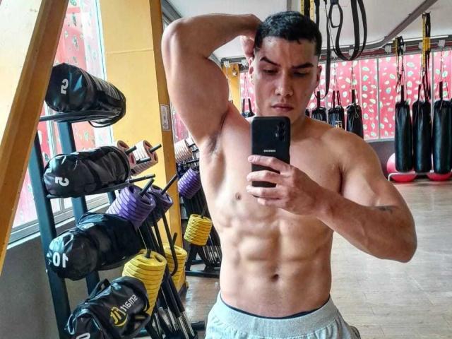 JuanJsmith, shirtless, muscular with defined abs, holding phone, taking mirror selfie, right arm raised, gray shorts, gym, ropes, weights in background.