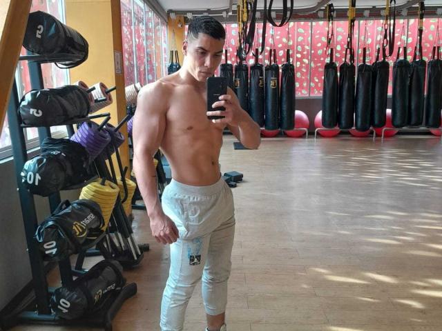 Muscle definition on JuanJsmith's bare chest, wearing gray sweatpants, standing in gym, taking mirror selfie with phone, holding black device, looking at camera, short dark hair, wooden floor.