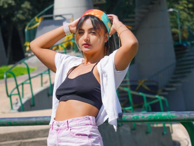 CloeesSuarez with hands in colorful cap, black sports bra, pink high-waisted shorts, white shirt, pink lips, white wristband, standing by green railing, looking confident, midday sunlight.