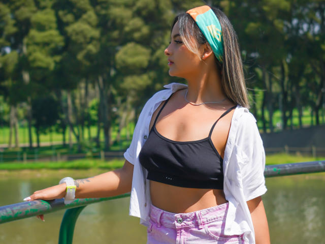 CloeesSuarez, met een bruine huid, een zwarte sportbeha, roze jeans, een wit shirt, een groene hoofdband en een polshorloge, leunt tegen een groene reling en laat haar kleine borsten en getoonde billen zien..