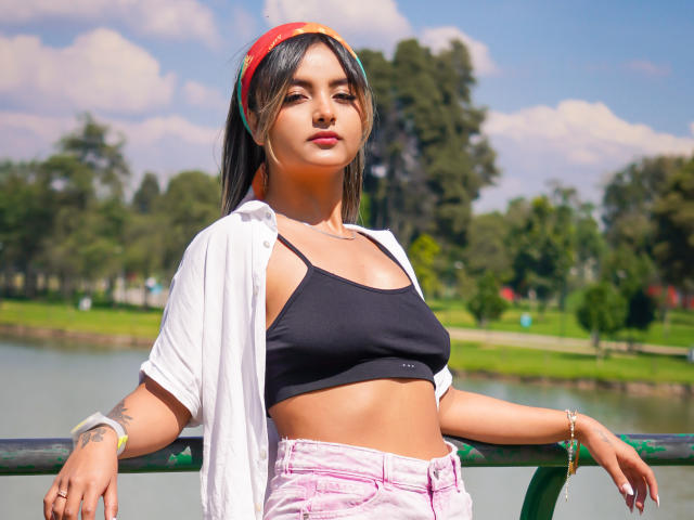CloeesSuarez with small tits, black sports bra, white shirt, pink jeans, colorful headband, tattoos, bracelet, leaning on green railing, sunny park, blue sky, fluffy clouds.