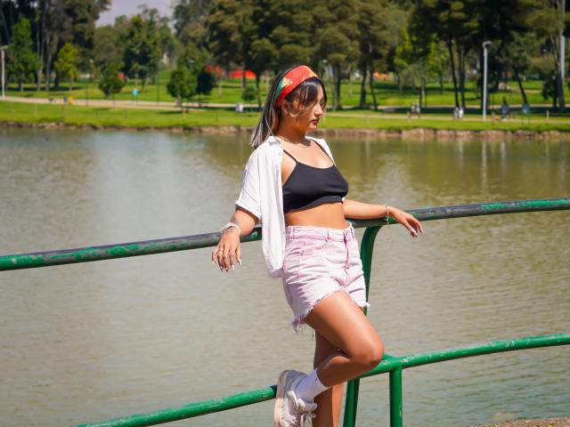CloeesSuarez leans on a green railing, wearing a black sports bra, pink shorts, white socks, and sneakers, with a red headband, by a pond, showing her ass.