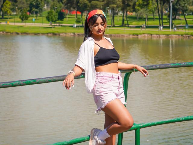 CloeesSuarez with tan skin, black bra, pink shorts, white socks, and cap, leaning on green railing by a pond, wearing open white shirt, showing small tits and ass.