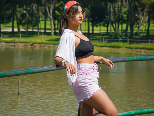 CloeesSuarez with medium tits, black sports bra, pink frayed shorts, white shirt, red headband, leaning on green railing by a pond, tattoo on left forearm, sunny park.