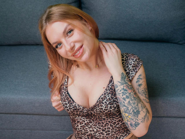 SlaviaH-hot with blonde hair, smiling, wearing a leopard print dress, showing her large tits, left arm tattooed, grey sofa behind, right hand touching her neck.