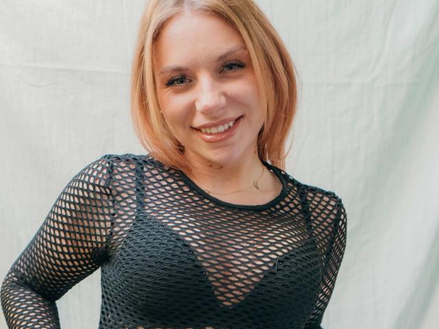 SlaviaH-hot has blonde hair, smiling, and is wearing a black fishnet top that reveals her tits. She's standing against a white background, looking cute and sexy.