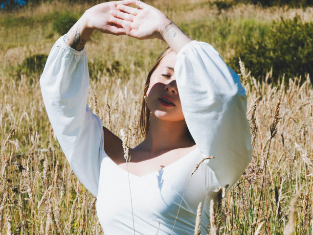Natsoriam with arms raised, wearing a white off-shoulder dress with puffy sleeves, standing in a wheat field, sunlight on her face, small tattoo on her right wrist.
