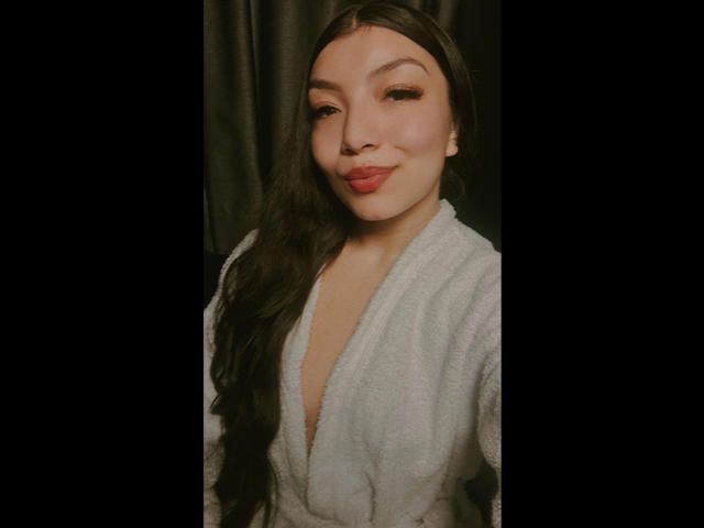 IbbyeMoreau with long black hair, pouty pink lips, and a light gray, deep V-neck, fluffy robe, smiling slightly, black background.