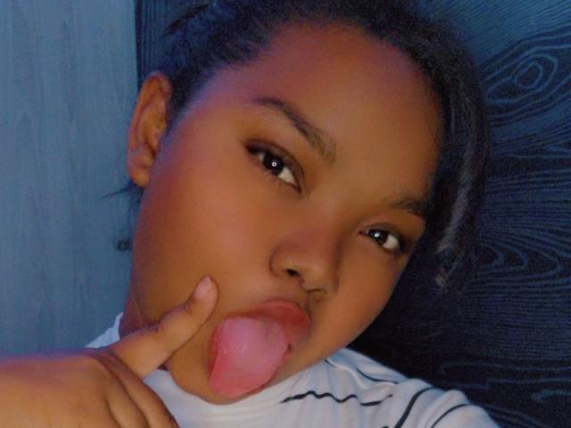 HotLindah with dark skin, black hair, and brown eyes, sticking out her pink tongue while touching her lips with her finger, wearing a white striped shirt.