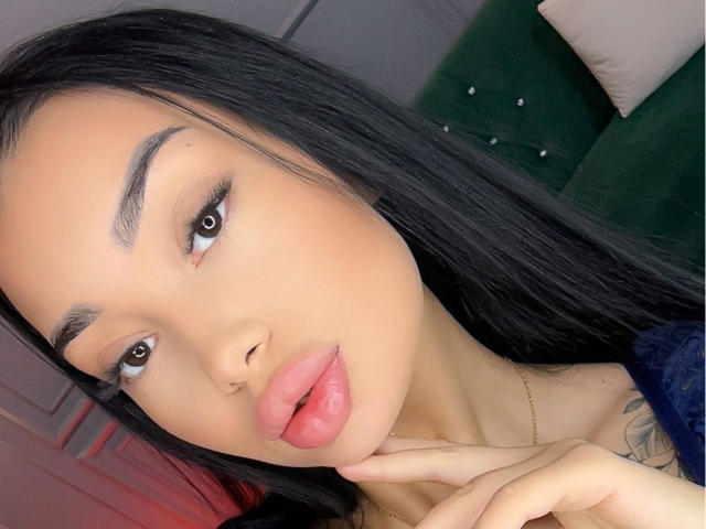 SashaFerrera with long black hair, full pink lips, and brown eyes, touching her chin, wearing a dark blue top, showing a tattoo on her right shoulder.