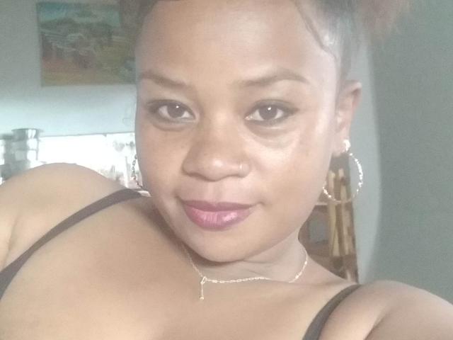 Danyiana69 has brown skin, purple lipstick, small nose ring, hoop earrings, and a thin necklace. She's wearing a black strap top, showing her bare shoulders and upper chest.