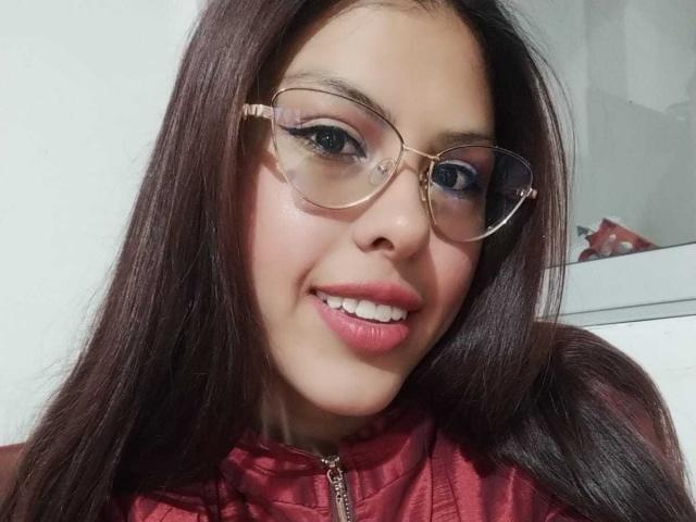 ElaiaaBakerr has long brown hair, wearing glasses, and a red zip-up shirt, with pink lipstick and a smile showing her teeth. Her ass is not visible.