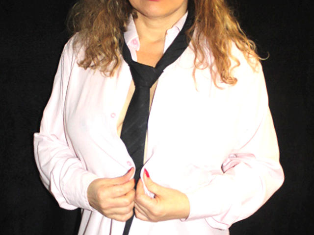 PriscillaHotMature, wearing an open pink shirt with a black tie, has curly brown hair, and is holding the tie with both hands, exposing her small tits.