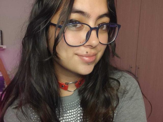 PerfectAnabelle with dark hair, blue glasses, nose piercing, lip piercing, red collar, gray shirt, smiling, looking sideways. Boobless, no visible ass or pussy. Medium skin tone, cute.