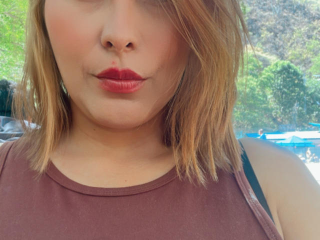 Het gezicht van SaritaVelez, met rode lippenstift, lichtjes opgetrokken lippen, lichte huid, blond haar tot op de schouders, draagt een bruin topje zonder mouwen, een zwarte band is zichtbaar aan de rechter schouder..