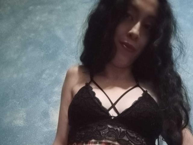 KendraSimons with long black hair, wearing a black lace bralette that shows her small tits and crisscross straps, has a subtle smile, and is standing against a textured blue background.