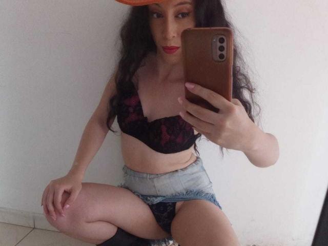 KendraSimons with long black hair, red lipstick, black lace bra, and denim shorts, squatting, taking a mirror selfie with an iPhone, showing her tits and pussy.
