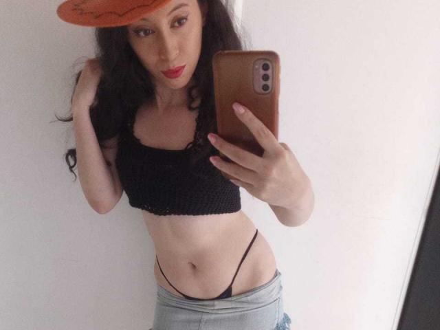 KendraSimons, com cabelos longos e escuros, vestindo uma blusa cropped preta e calças cinzas puxadas para baixo para revelar uma tanga preta, segurando um celular laranja, selfie no espelho, lábios vermelhos, parede branca..