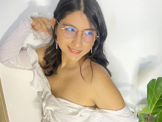 Eine Frau mit langen, schwarzen Haaren, Brille, einem weißen Oberteil mit einem Schulterteil, die lächelt, ihr Ohr berührt, ihre nackte Schulter zeigt, eine Zimmerpflanze mit grünen Blättern auf der rechten Seite, weißer Hintergrund, warmes Licht..