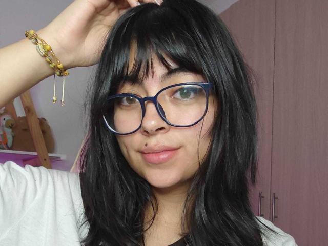 PerfectAnabelle with black hair, blue glasses, and a nose ring, wearing a white shirt, and a gold bracelet, smiling at the camera. Her green eyes are slightly open.