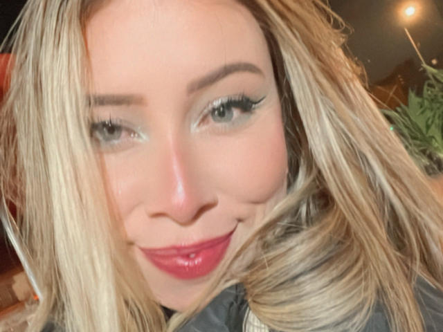 JennyLawry has blonde hair, green eyes, and is smiling with pink lipstick. Her eyelashes are long and she looks cute. Her tits are not visible in this shot.