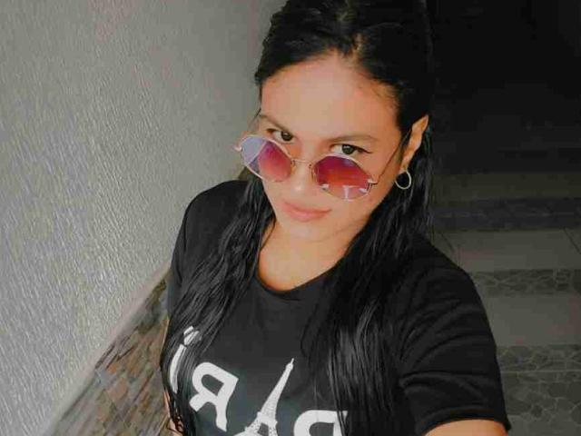 SweetAlma69 with dark hair, wearing a black T-shirt, pink-tinted sunglasses, and hoop earrings, smiling slightly, standing against a textured wall, in a dimly lit stairway.