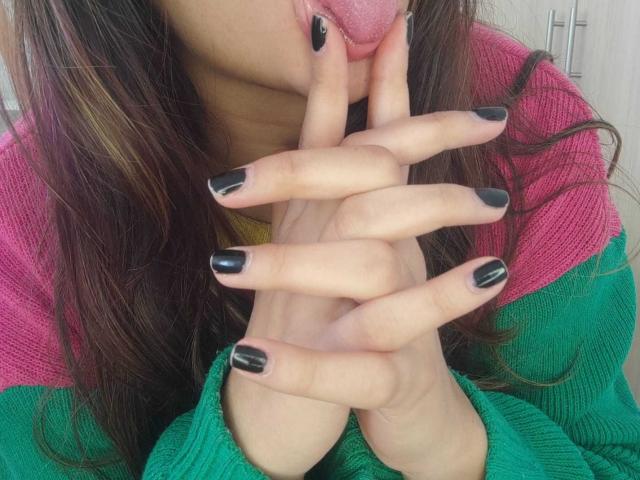 PerfectAnabelle's tongue is out, licking her black-manicured fingers. She's wearing a green and pink sweater, with her brown hair slightly visible. Her fingers are stacked, tongue out.