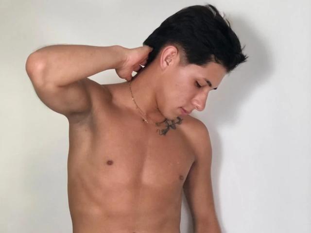 SoffiaVegga, shirtless, with a tattoo on the left side of his neck, has short black hair and a gold necklace, touching his right ear, with a relaxed, downward gaze.