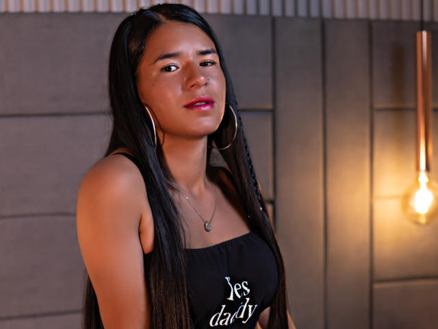 AliceRider has long black hair, red lipstick, hoop earrings, and a black tank top showing "yes daddy" in white. She has a cute, sexy face and a small nose.