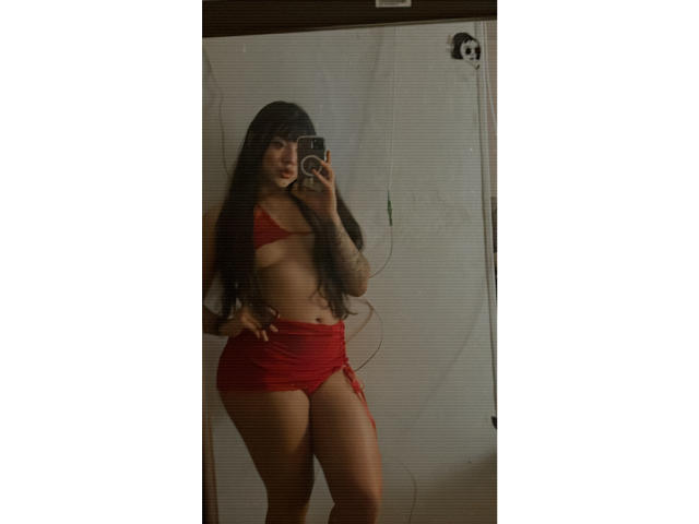 SteffanyTaylor with long dark hair, red bikini top showing small tits, red shorts with a side tie, tattoo on right arm, taking a mirror selfie, holding phone in left hand.