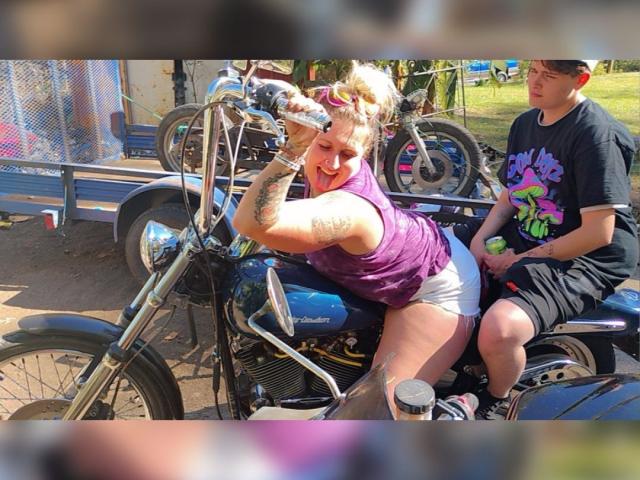 Eine Frau namens HeavyChevy, mit Tattoos, einem lila Tanktop und weißen Shorts, trinkt aus einer Flasche und lehnt sich an ein blaues Motorrad, während ein Mann in einem schwarzen Hemd hinter ihr sitzt..