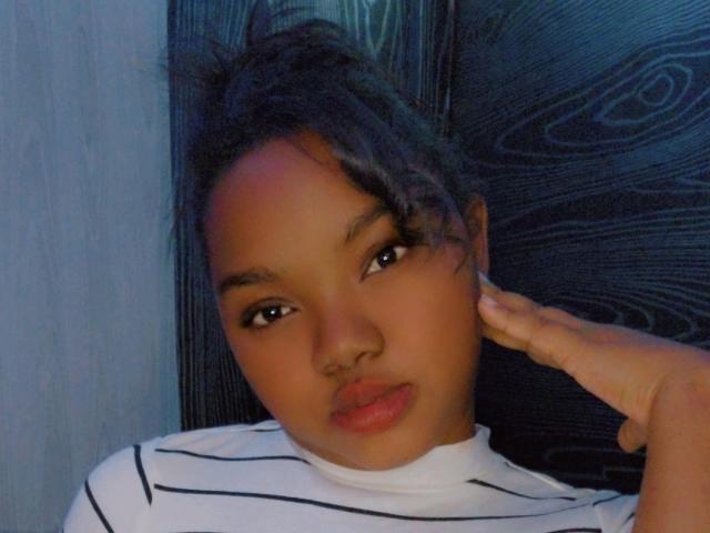 HotLindah with brown skin, dark hair in a messy bun, wearing a white striped shirt, has a hand on her cheek, looking at the camera with pouty lips.