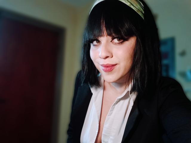 SpicyGrace, con un caschetto nero, una fascia verde, un blazer nero, una camicia bianca, labbra carnose e occhi marroni, guarda seducentemente la fotocamera, mostrando leggermente il seno attraverso la camicia aperta..