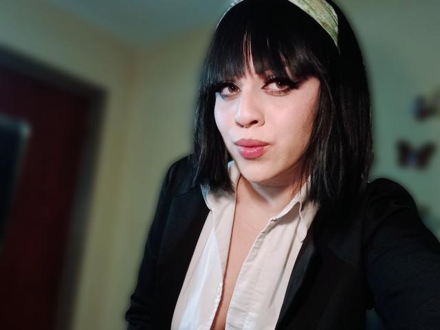 SpicyGrace ha i capelli neri tagliati a caschetto, una fascia verde, un blazer nero, una camicia bianca, labbra piene e un trucco per gli occhi scuro, e guarda leggermente di lato con un'espressione sicura..
