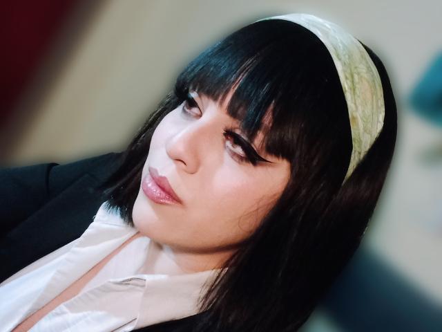 SpicyGrace ha i capelli neri, una fascia verde e guarda leggermente verso l'alto con un'espressione accigliata. Indossa un blazer nero sopra una camicia bianca..