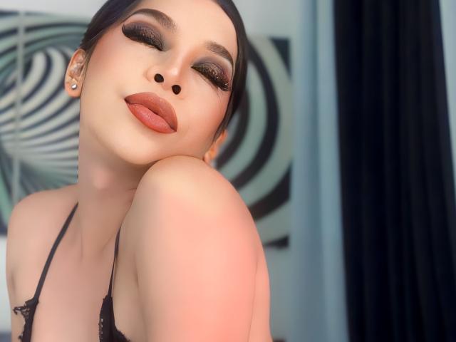 NattiStarr with her eyes closed, dark lipstick, and black eyeliner, head resting on her knee, wearing a black lace bra, looking sexy with her tongue slightly out.