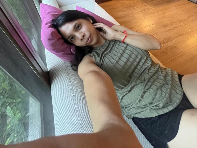 NattiStarr with dark hair, green ribbed sleeveless top, black shorts, pink pillow, red bracelet, wooden floor, taking selfie, smiling, hand on cheek, window with green plants.