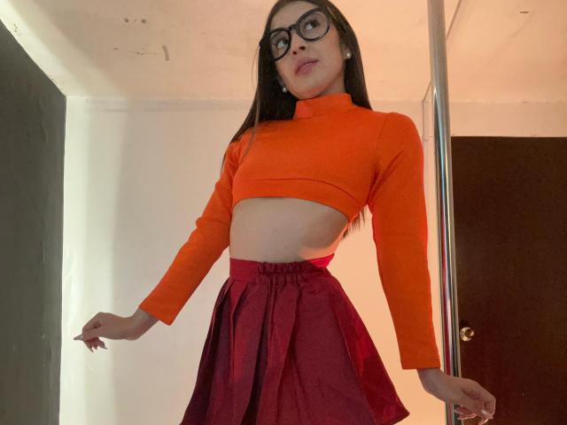 Emma Lombardi, com cabelos longos e pretos, óculos, cropped top laranja, saia vermelha de cintura alta, em pé ao lado de um poste prateado, exibindo um abdômen fino e plano, e uma expressão adorável e confiante..