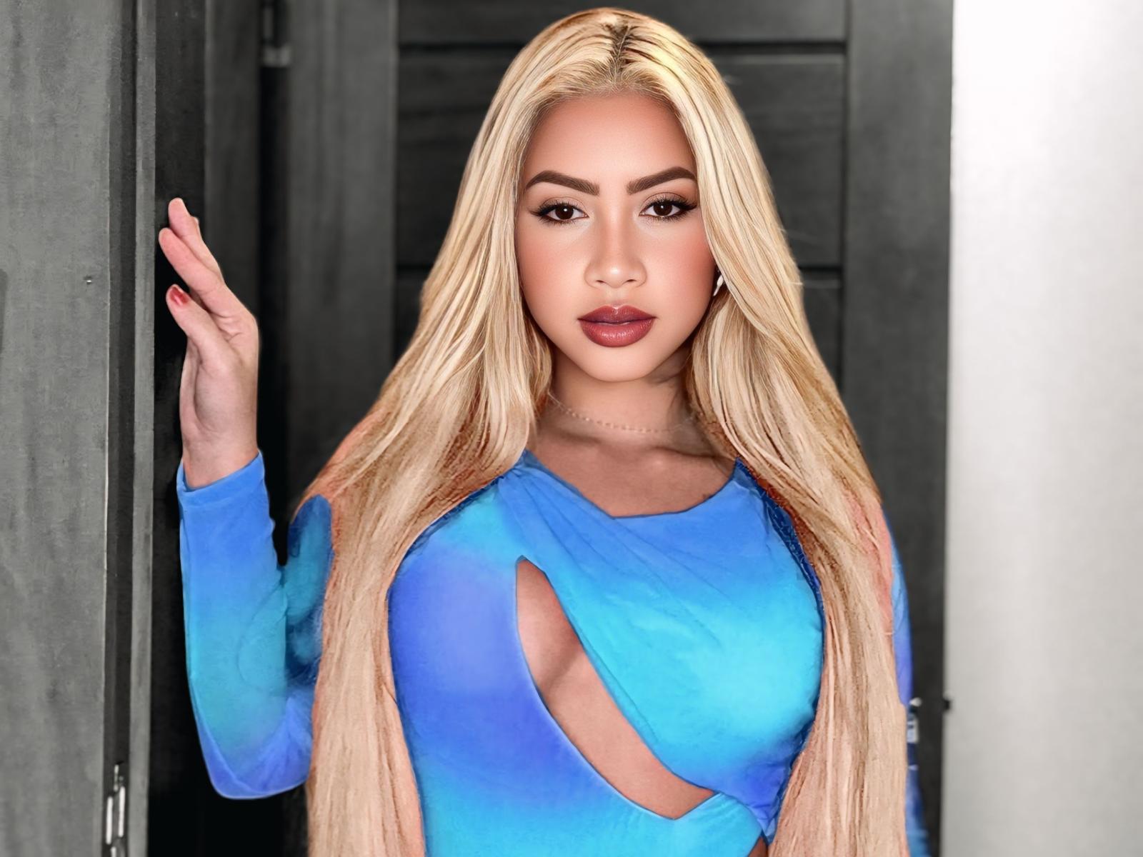 Blonde Yaraah with long hair, blue cut-out dress, large tits, red nails, and makeup, standing in doorway, looking at camera.