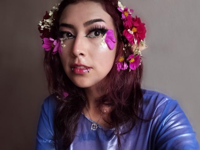 AlicePhantom with dark red hair, wearing a blue shiny top, flower crown, pink flower on cheek, teardrop makeup, lip ring, and necklace, looking up with sad eyes.