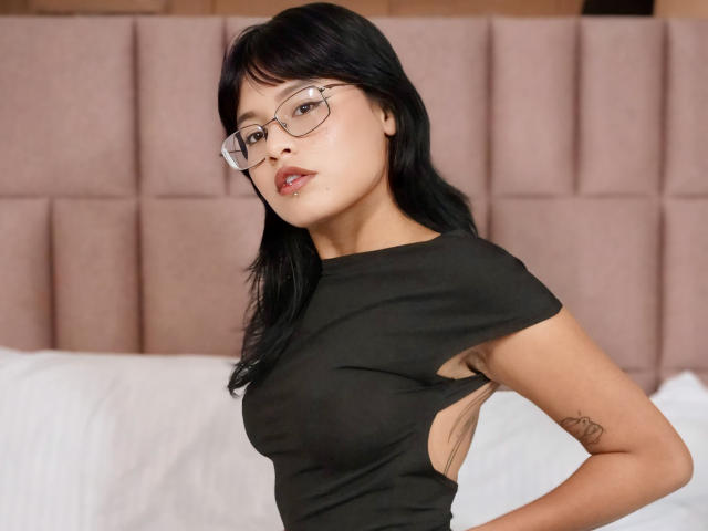 ElizabethRoux with black hair, glasses, and a black shirt showing her sideboob, arms raised, tattoo on her right arm, in a bedroom with a pink upholstered headboard.