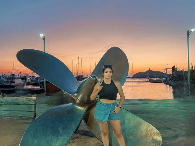 NovaDixon stands in front of a metal boat sculpture, wearing a black sports bra and blue shorts, with a sunset and docked boats in the background.