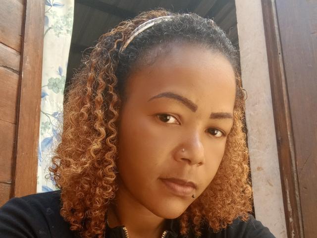 Araf has curly, light brown hair with a white headband, brown skin, a nose piercing, and a lip piercing, wearing a black top, looking slightly to the side.