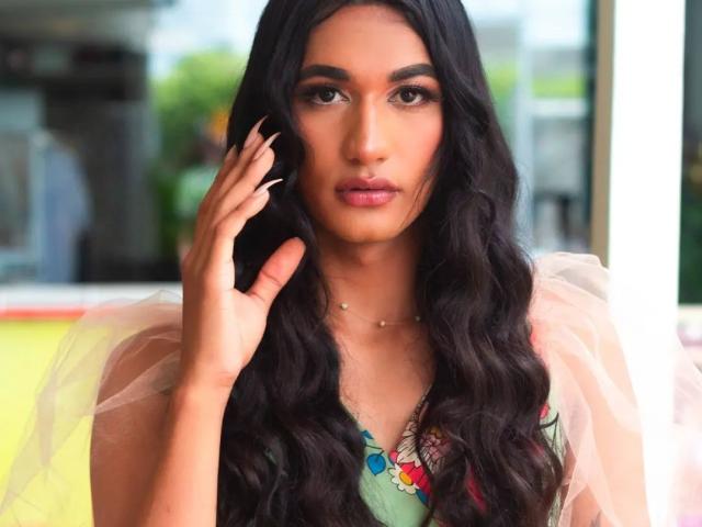 Alexa69s with long black wavy hair, brown eyes, and pink lipstick, wearing a green dress with colorful embroidery, and a pink tulle shoulder, touching her hair.