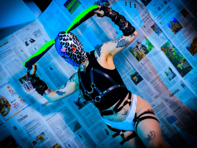 AmySpider with leopard mask, black harness, white panties, black fingerless gloves, neon green belt, and tattoos, posing with arms raised, showing a tattooed ass and white panties.